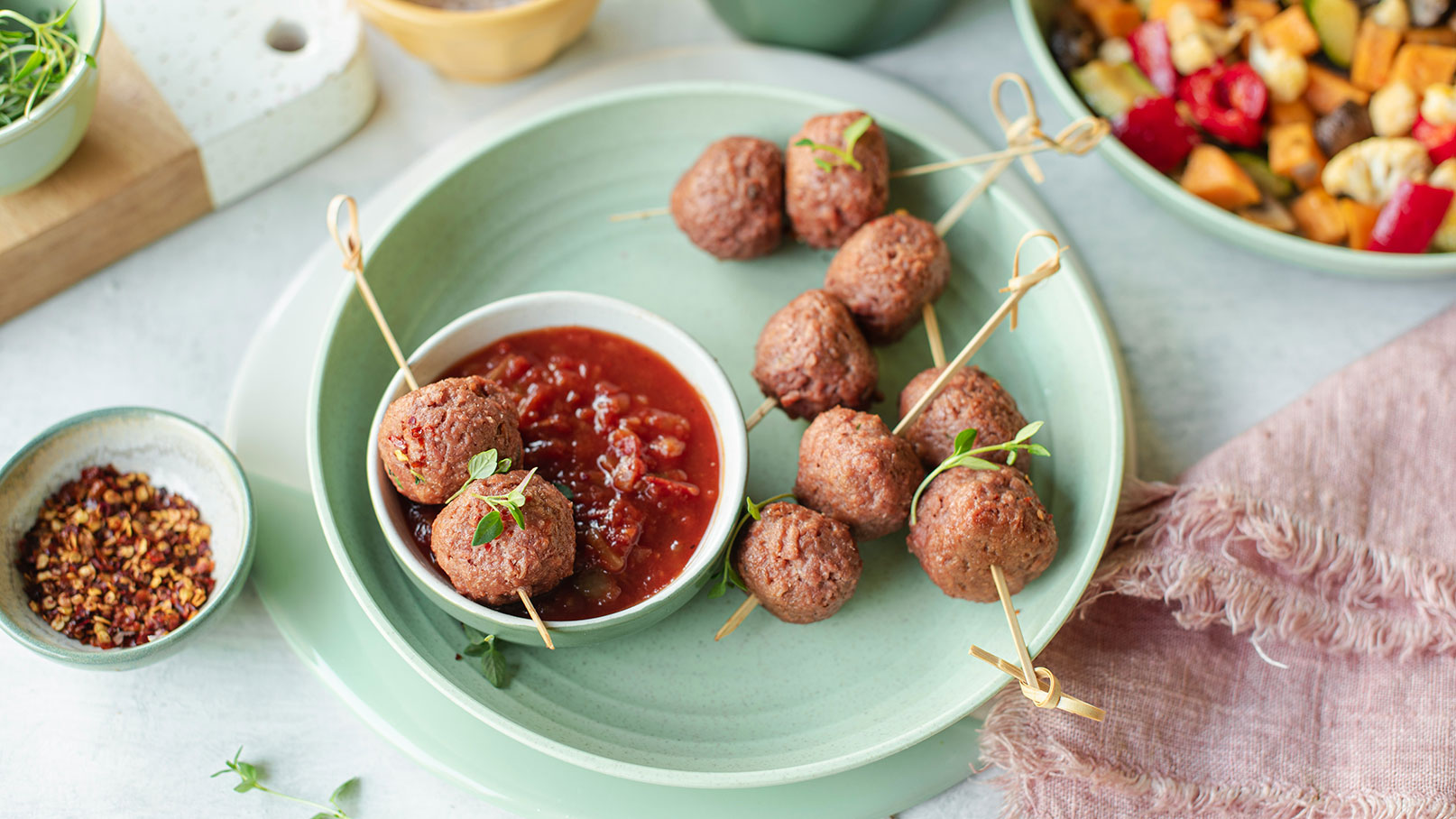 Eine grüne Servierplatte mit Fleischbällchen auf Spießen und einer kleinen Schüssel mit Tomatensoße. Daneben liegen weitere Fleischbällchen und ein kleiner Schüssel mit roten Chili-Flocken. Im Hintergrund ist ein bunter Gemüsesalat sichtbar.