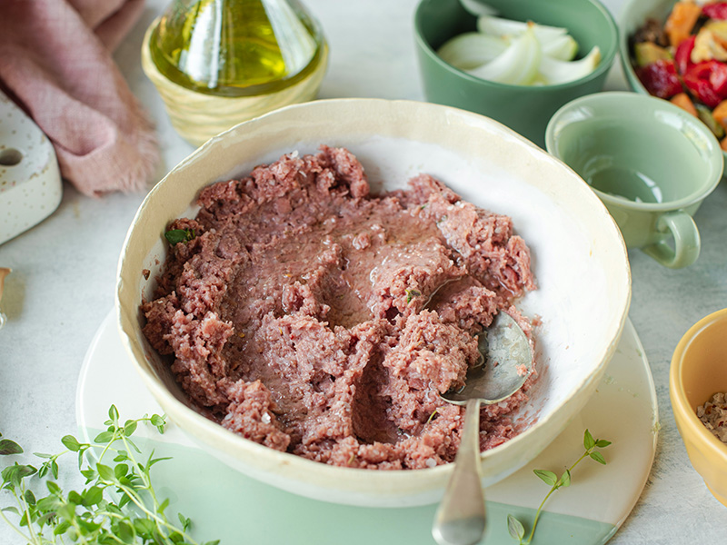 Eine Schüssel mit grob püriertem Fleisch, umgeben von Kräutern. Neben der Schüssel sind verschiedene Zutaten wie Zwiebeln, Öl und kleine Schalen mit weiteren Lebensmitteln zu sehen. Die Textur des Fleisches ist dick und feucht.