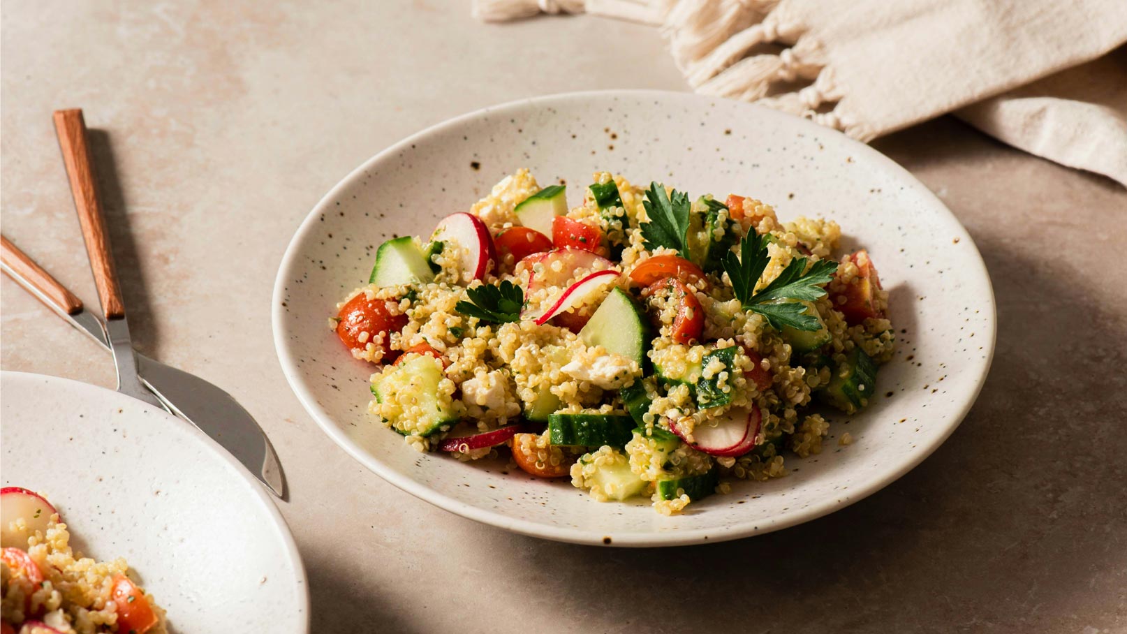 Eine Schüssel Salat mit Quinoa, frischen Gemüsestücken von Gurke, Tomate und Radieschen sowie frischen Kräutern. Der Salat ist bunt und ansprechend angerichtet, und die Schüssel steht auf einer neutralen Oberfläche.