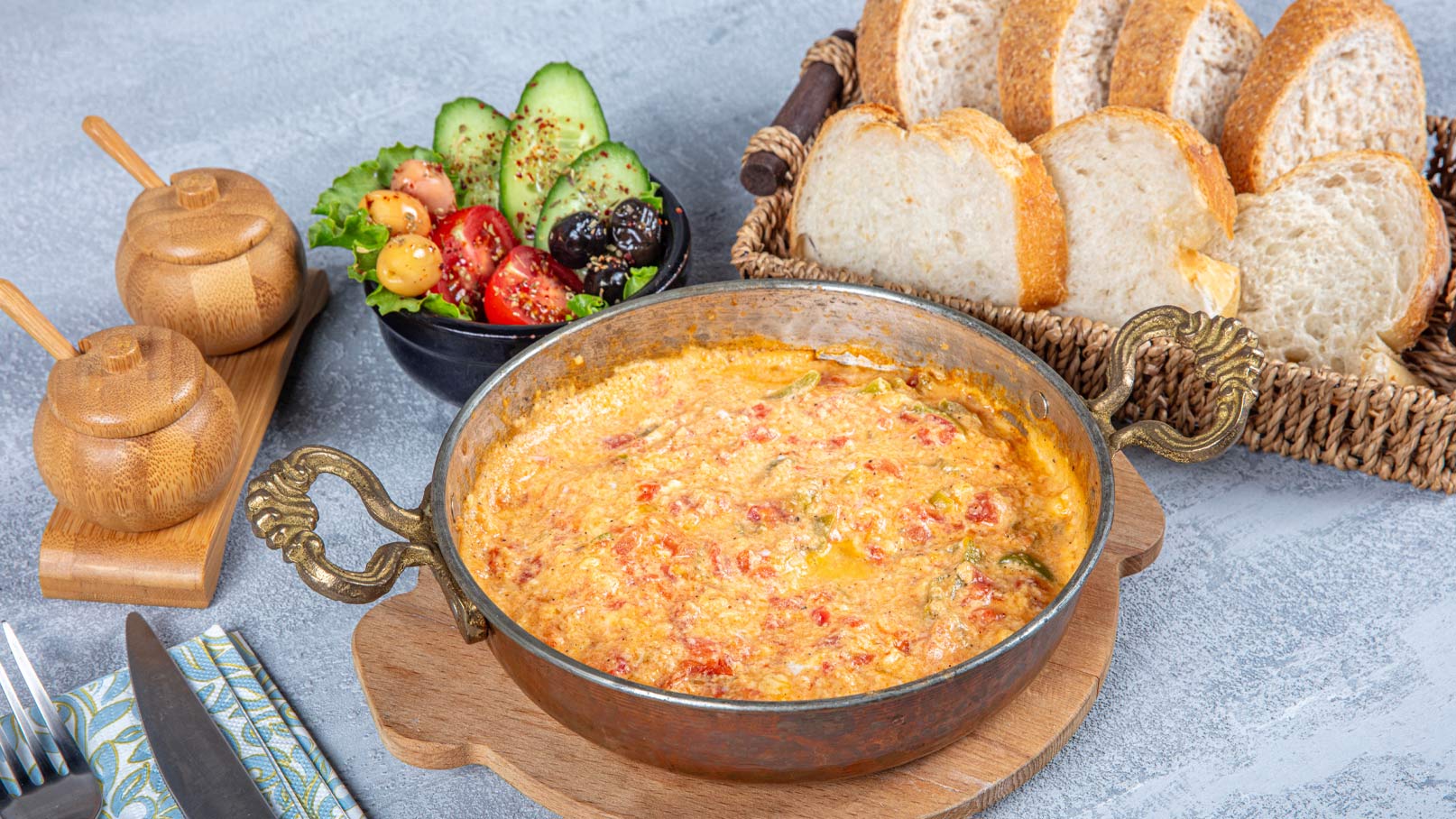 Ein rustikales Gericht in einer bronzefarbenen Pfanne, umgeben von frischem Brot. Daneben ein Salat mit Gurken, Tomaten und Oliven in einer schwarzen Schüssel. Zwei kleine, holzfarbene Behälter stehen auf einem Holzbrett. Der gesamte Tisch ist appetitlich gedeckt.