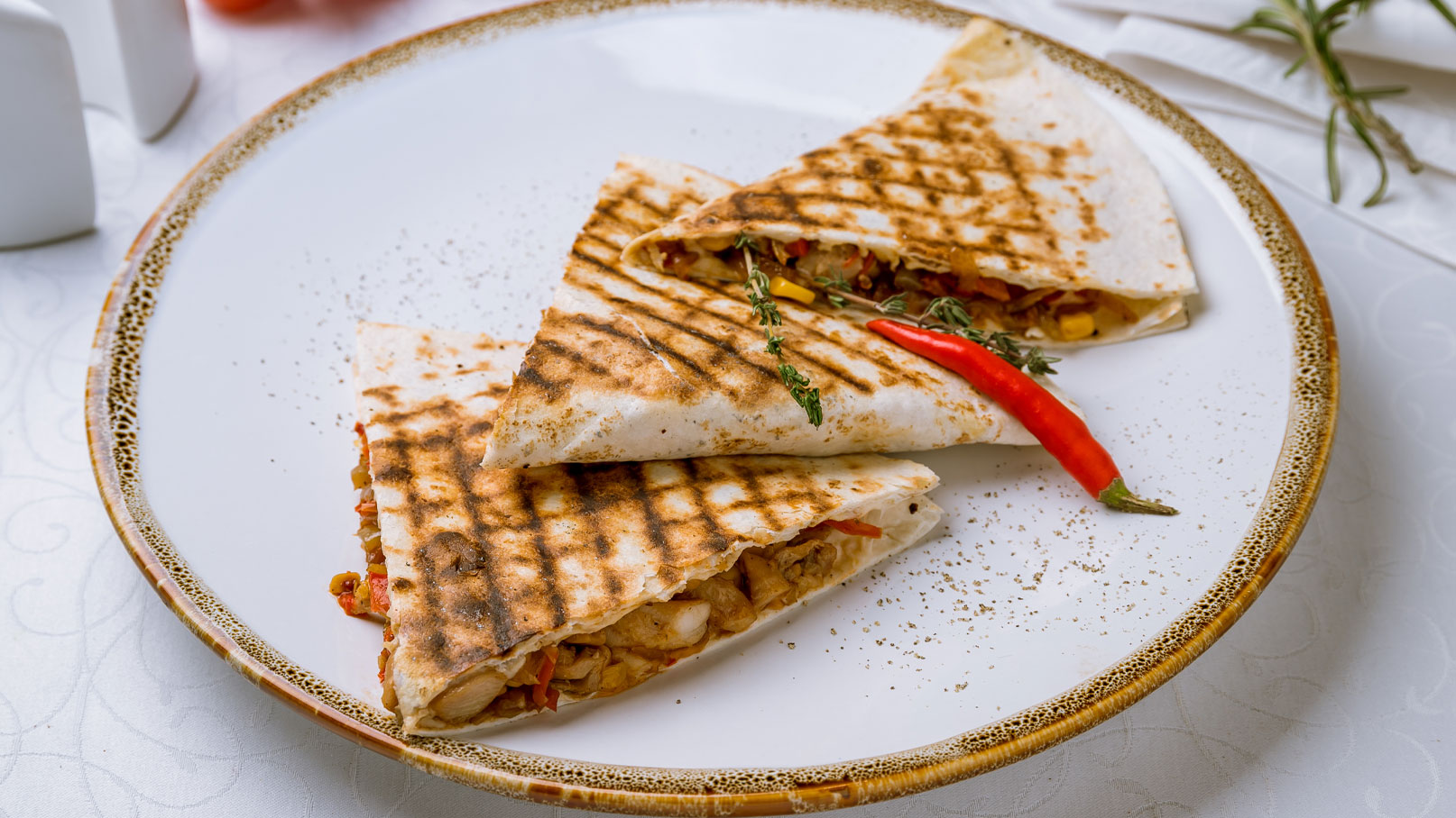 Auf dem Bild sind zwei gegrillte Tortillas auf einem weißen Teller zu sehen. Die Tortillas sind halbiert und gefüllt mit buntem Gemüse und Hähnchen. Eine rote Chili und einige Kräuter sind dekorativ darauf platziert.