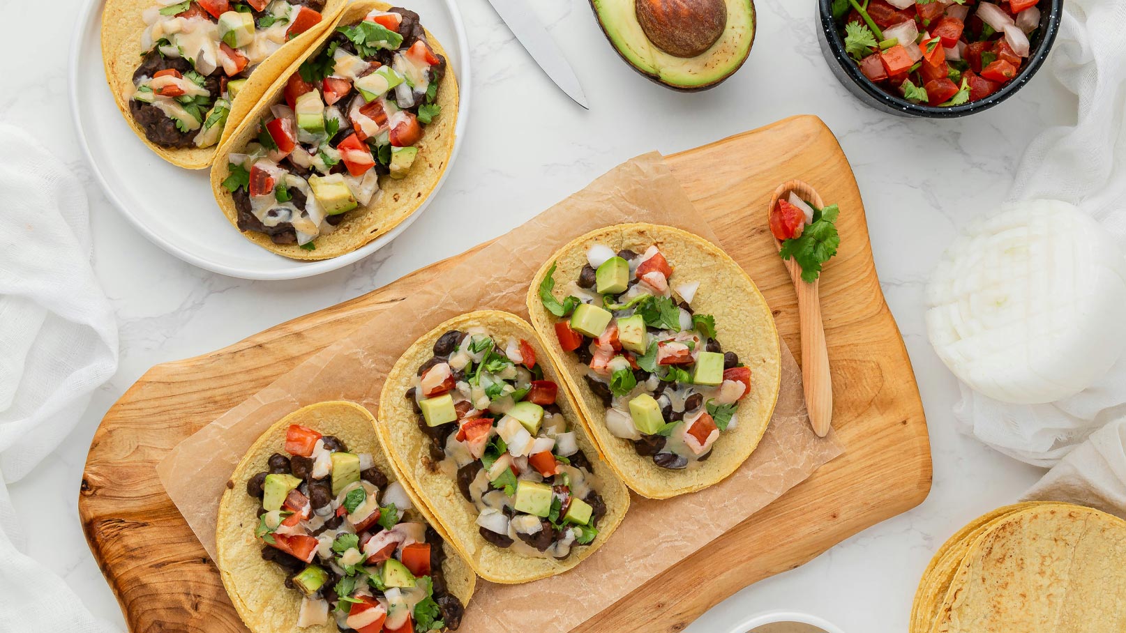 Drei gefüllte Tacos liegen auf einer Holzplatte. Sie sind mit schwarzen Bohnen, frischem Gemüse, Avocado und Koriander belegt. Daneben befindet sich eine Schüssel mit Salsa und einige Tortillas.