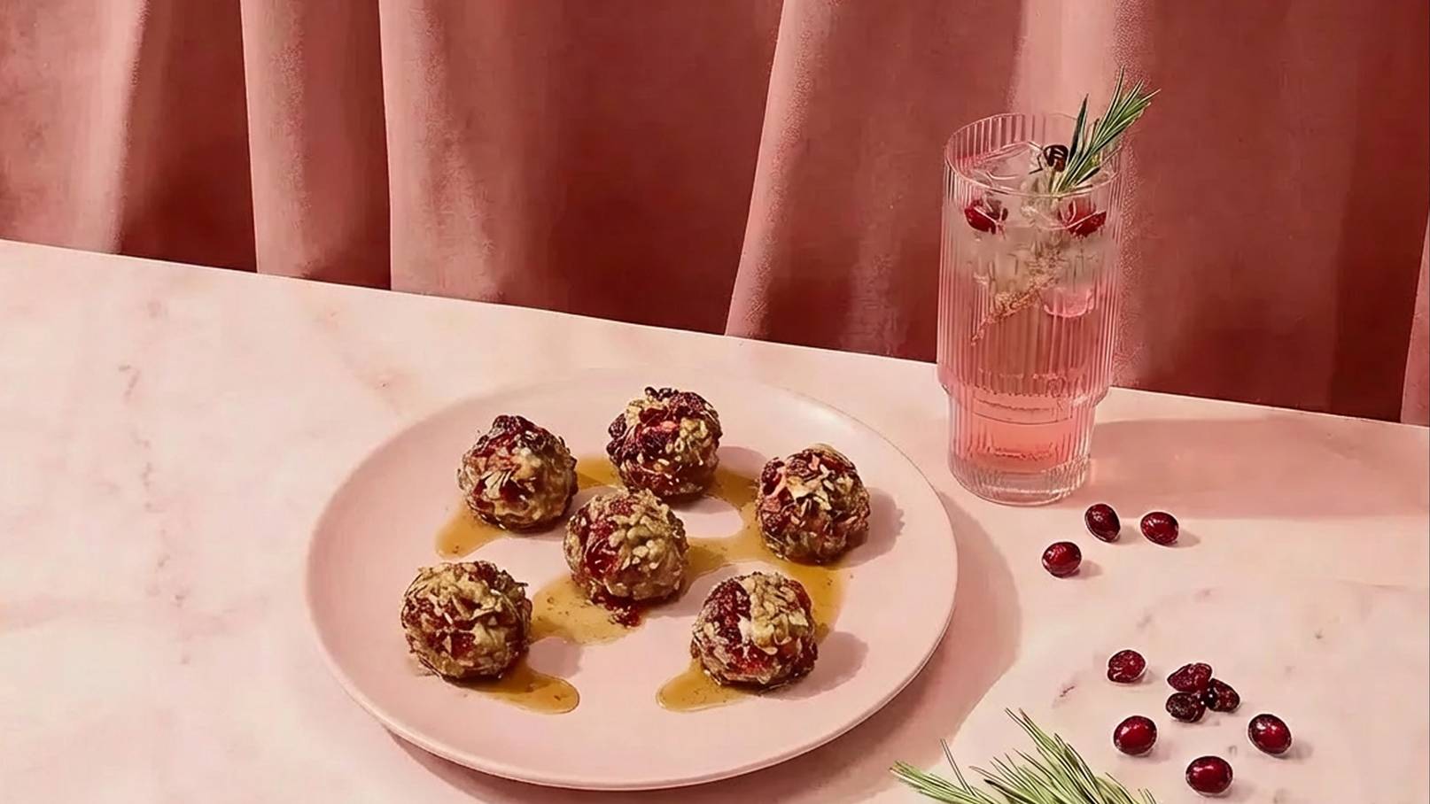 Sechs energiebällchen in einer Schale, umgeben von einem süßen Sirup. Daneben steht ein Glas mit rosa Drink und Dekoration aus Kräutern und Granatapfelkernen. Der Hintergrund ist in sanften Rosatönen gehalten.