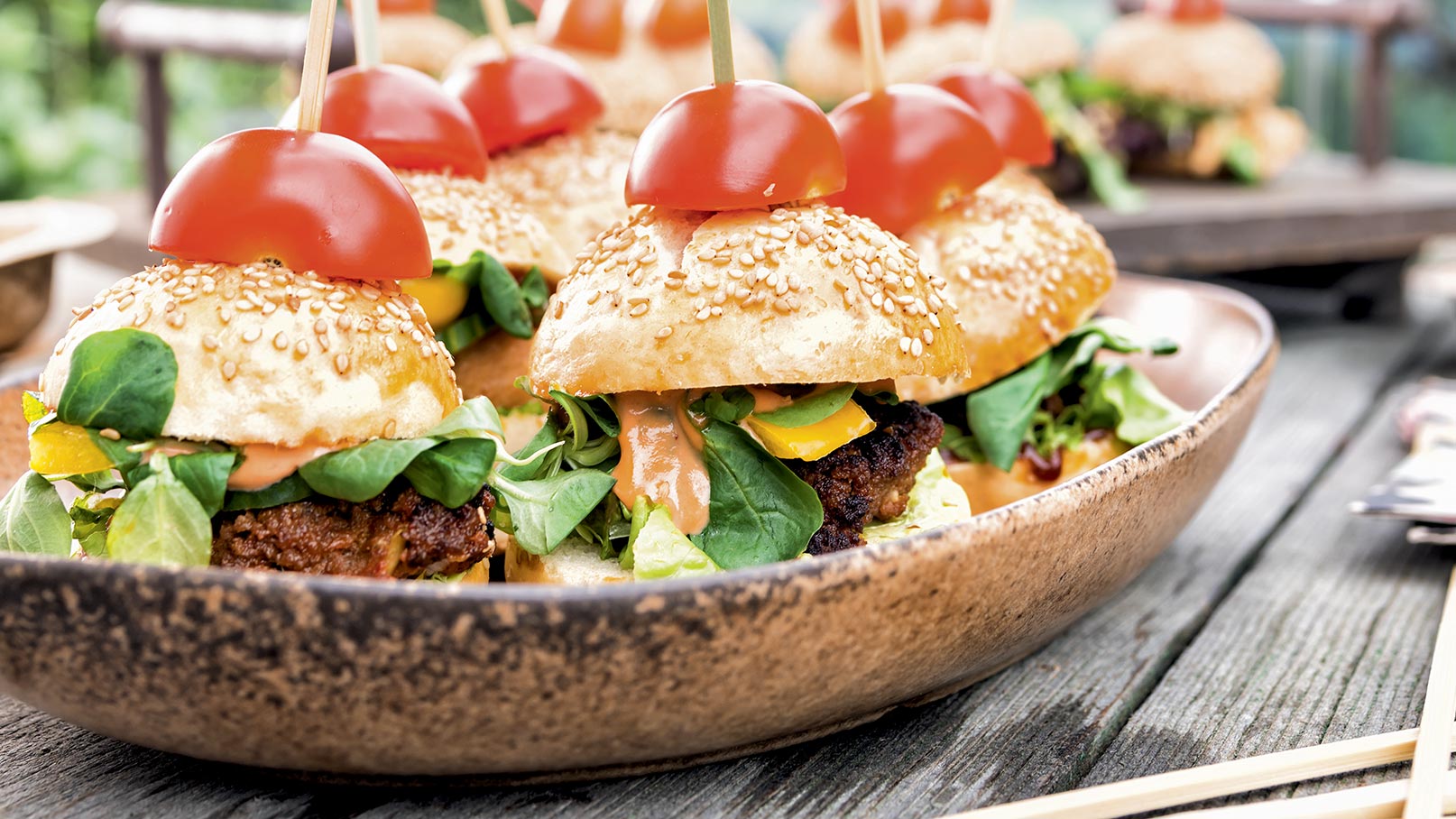 Eine Schüssel mit Mini-Burgern, die mit Sesambrötchen, frischem Spinat, Paprika und einer Sauce gefüllt sind. Auf jedem Burger befindet sich eine halbierte Kirschtomate, die auf einem Holzspieß steckt. Der Hintergrund zeigt eine rustikale Tischoberfläche.