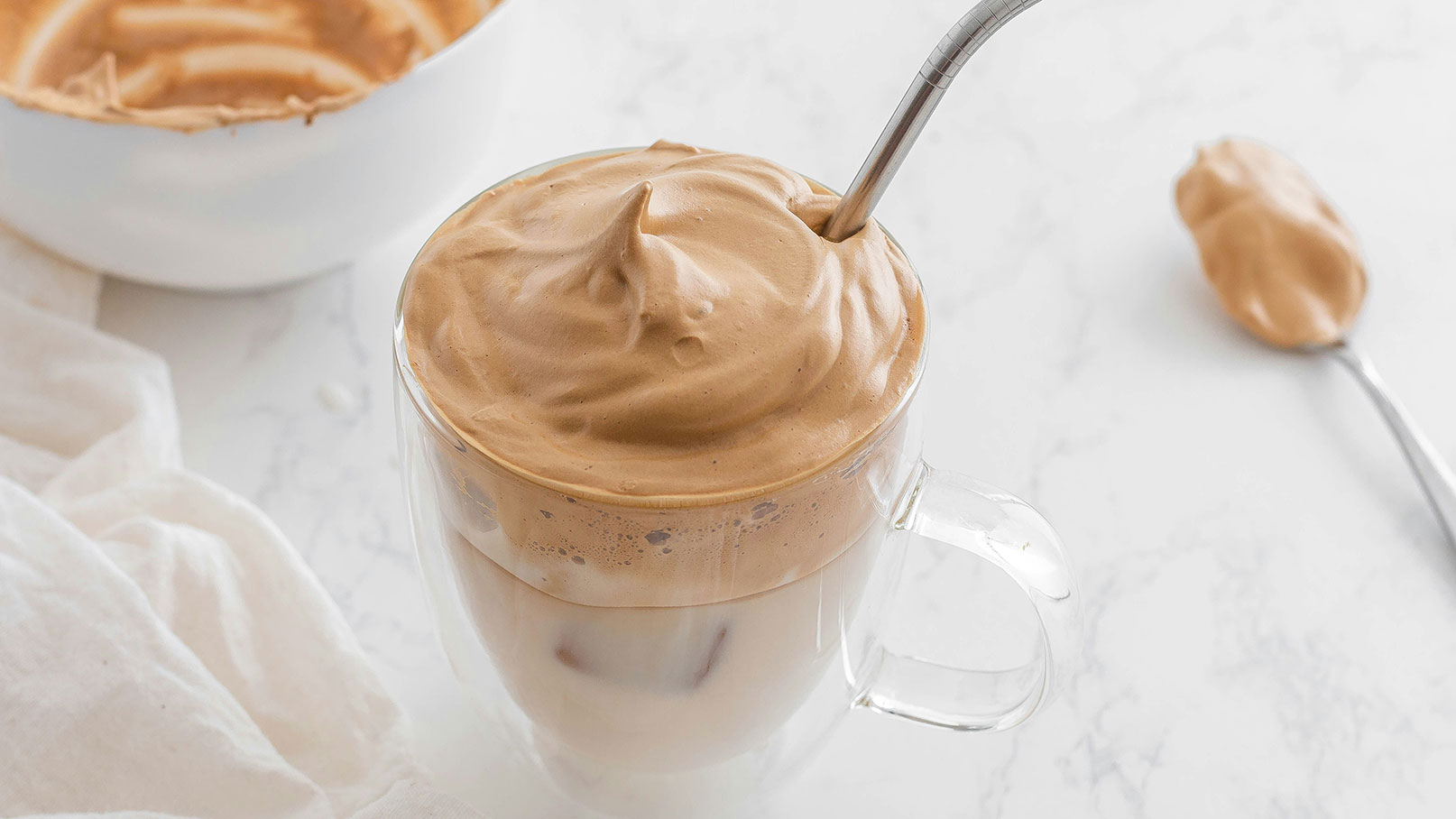 Ein transparentes Glas enthält einen cremigen, schaumigen Kaffeegenuss mit einem Löffel Eiskaffee. Der Kaffee hat eine hellbraune Farbe und sieht luftig aus. Im Hintergrund ist eine Schüssel mit ähnlich gefärbtem Inhalt zu sehen.