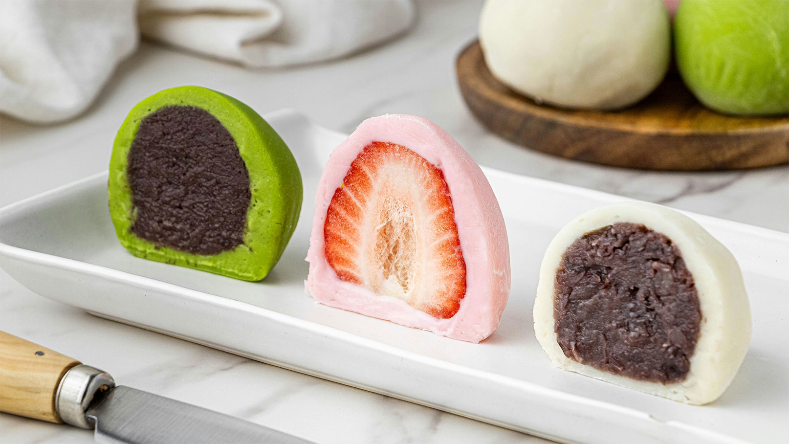 Drei halbe Mochis auf einem weißen Teller: ein grüner mit einer Füllung aus roter Bohnenpaste, ein rosa mit einer Erdbeerfüllung und ein weißer ebenfalls mit roter Bohnenpaste. Im Hintergrund sind weitere, runde Mochis sichtbar.
