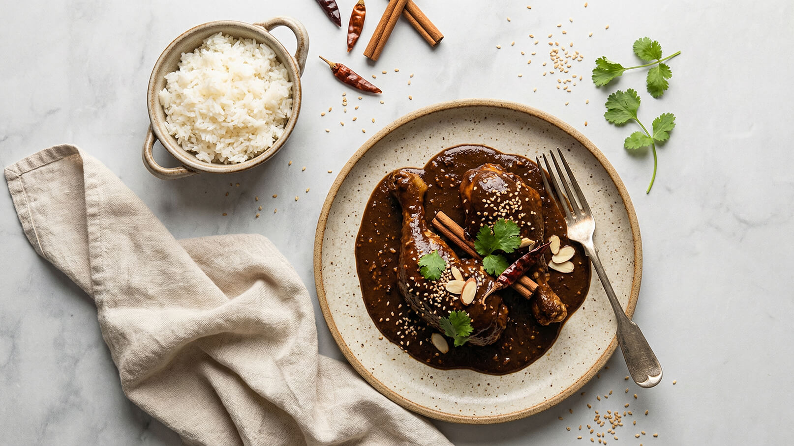Ein Teller mit zwei Hähnchenschenkeln in dunkler Sauce, dekoriert mit Sesam, frischem Koriander und Zimtstangen. Neben dem Teller befindet sich eine Schüssel mit gedämpftem Reis. Im Hintergrund sind einige getrocknete Chilischoten sichtbar.
