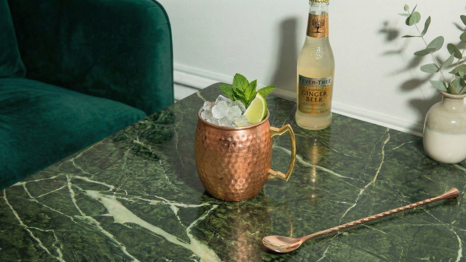 Auf einem marmorierten Tisch steht ein Kupferbecher gefüllt mit Eis, Minze und einer Limettenscheibe. Neben dem Becher steht eine Flasche Ginger Beer. Im Hintergrund ist ein grünes Sofa sichtbar.