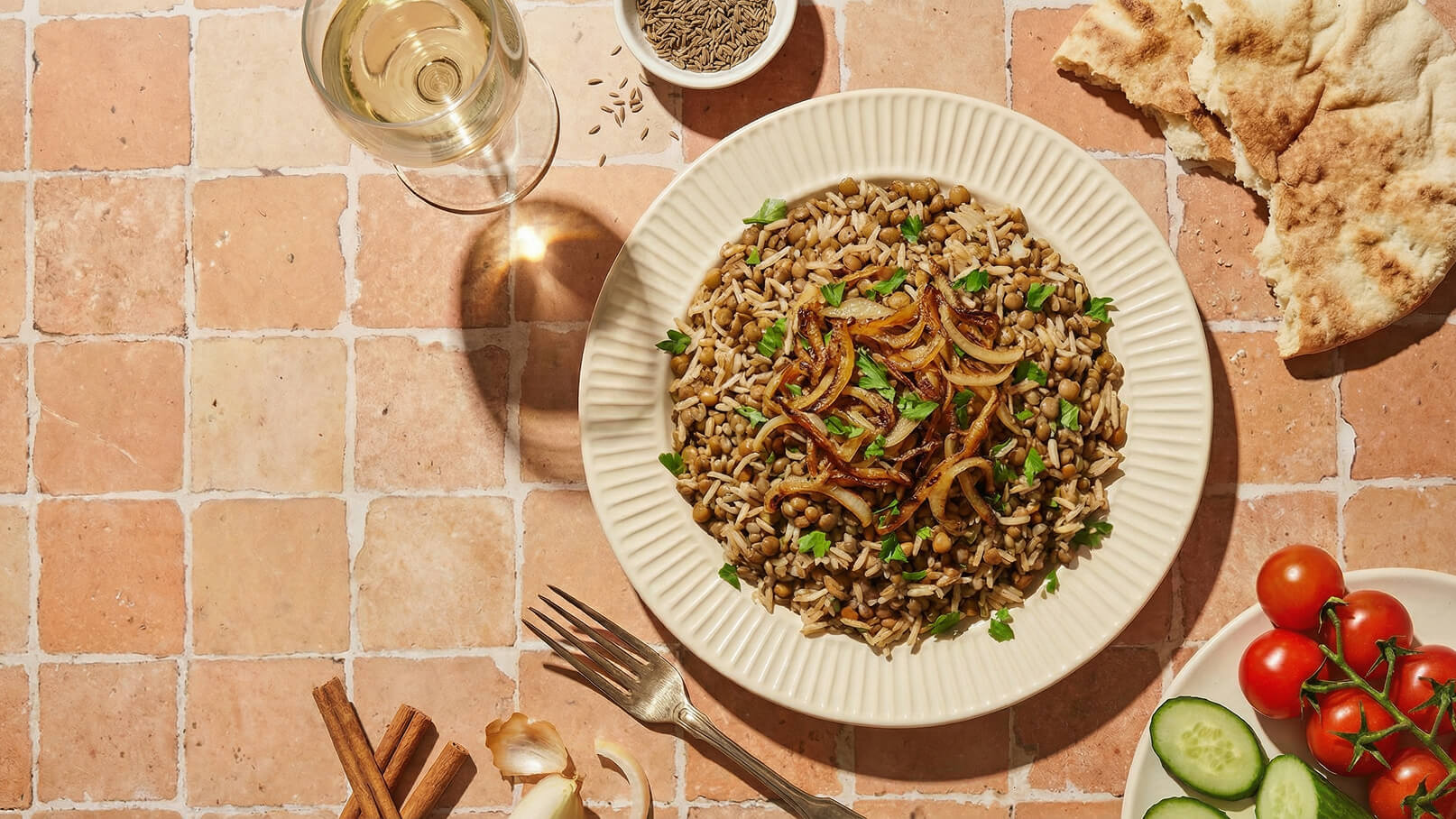 Ein Teller mit Reis und Linsen, garniert mit gebratenen Zwiebeln und frischer Petersilie, steht auf einem Tisch. Daneben liegt ein Stück Fladenbrot und eine kleine Schale mit Gewürzen. Auf der anderen Seite sind frisches Gemüse, darunter Tomaten und Gurken.