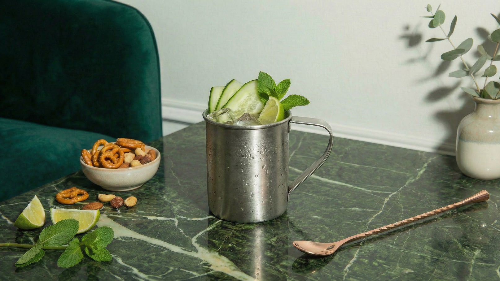 Ein metallischer Becher mit eisgekühltem Getränk, garniert mit Gurkenscheiben und Minzblättern, steht auf einem marmorierten Tisch. Daneben liegt eine kleine Schale mit Brezeln und Nüssen sowie mehrere Limettenschnitze.