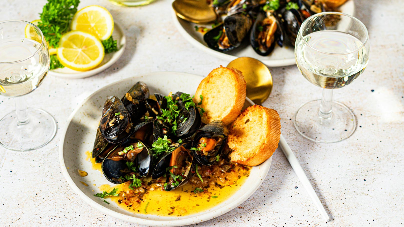 Ein Teller mit dampfenden Muscheln in einer würzigen Sauce, garniert mit frischem Koriander, dazu zwei Stücke geröstetes Brot. Im Hintergrund stehen Gläser mit Weißwein und frische Zitronenscheiben auf einem Teller.