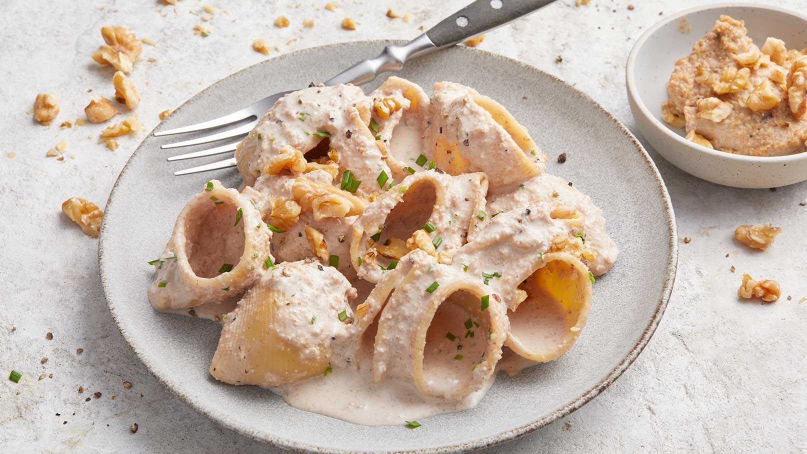 Ein Teller mit gefüllten Teigwaren in einer cremigen Sauce, garniert mit gehackten Walnüssen und frischen Kräutern. Daneben liegt eine kleine Schale mit einer ähnlichen Füllung. Eine Gabel befindet sich auf dem Teller.