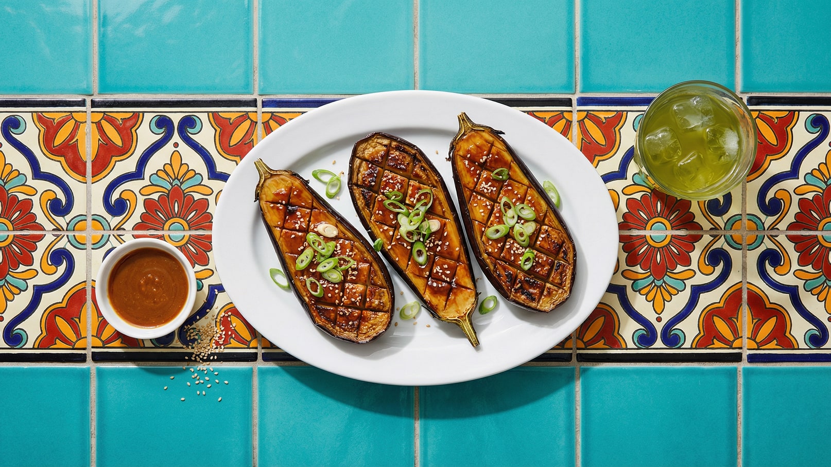 Ein Teller mit drei gegrillten Auberginenhälften, die mit einer glänzenden Sauce, Sesam und Frühlingszwiebeln garniert sind. Daneben ein kleiner Schüssel mit dunkler Soße und ein Glas mit einem grünen Getränk. Der Hintergrund besteht aus bunten Fliesen.