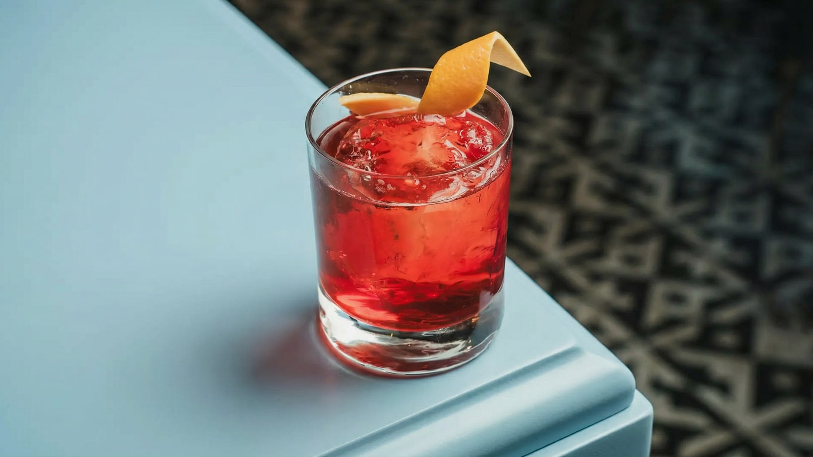 Ein Glas mit einem roten Getränk, gefüllt mit Eiswürfeln, steht auf einem blauen Tisch. Eine spiralförmige Kruste aus Zitrone liegt oben auf dem Getränk. Der Hintergrund zeigt ein geometrisches Muster.
