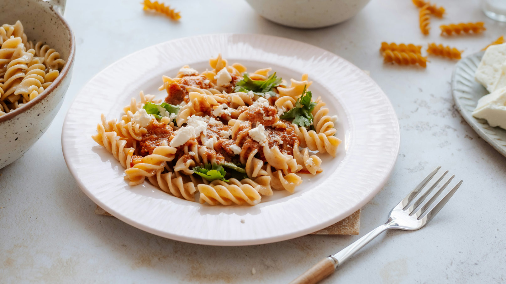 Ein Teller mit Spirali-Pasta, garniert mit einer Tomatensauce, frischen Kräutern und zerbröckeltem Feta-Käse. Im Hintergrund befindet sich eine Schüssel mit weiteren Pasta. Die Anordnung der Speisen wirkt appetitlich und einladend.