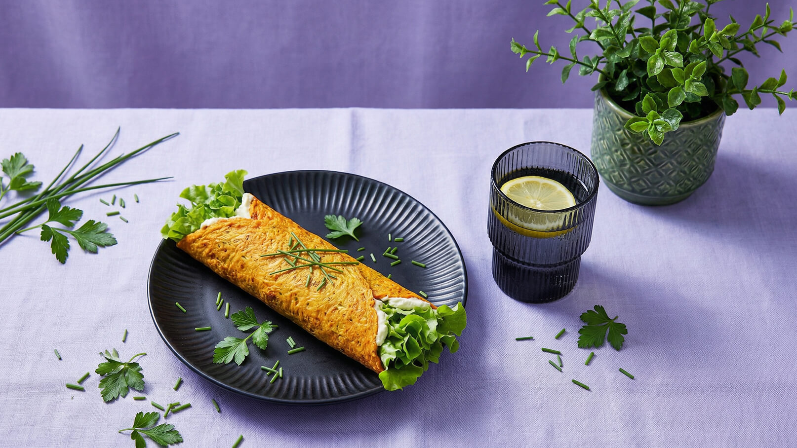 Auf einem schwarzen Teller liegt ein gefülltes Omelette mit frischem Salat und Kräutern. Daneben steht ein Glas mit Wasser und einer Zitronenscheibe. Im Hintergrund sind grüne Pflanzentöpfe und frische Kräuter verteilt. Der Tisch ist mit einem lila Tuch bedeckt.