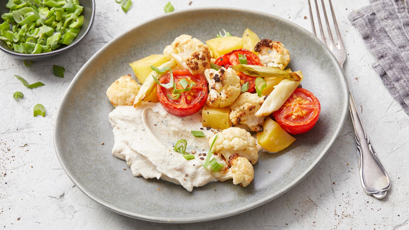 Eine graue Schüssel mit geröstetem Blumenkohl, Tomaten und Kartoffeln, garniert mit grünen Zwiebeln. Dazu serviert ist eine cremige Sauce. Die Farben sind lebendig und die Zutaten ansprechend angeordnet.