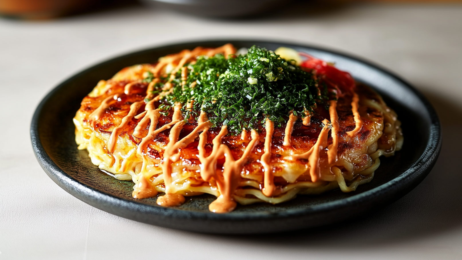 Ein schwarzer Teller mit einer Portion Okonomiyaki, einem japanischen Pfannkuchen. Der Pfannkuchen ist goldbraun gebraten und mit grünen Zwiebeln sowie einer hellen Sauce dekoriert. Gemüse ist sichtbar, und die Präsentation ist appetitlich.