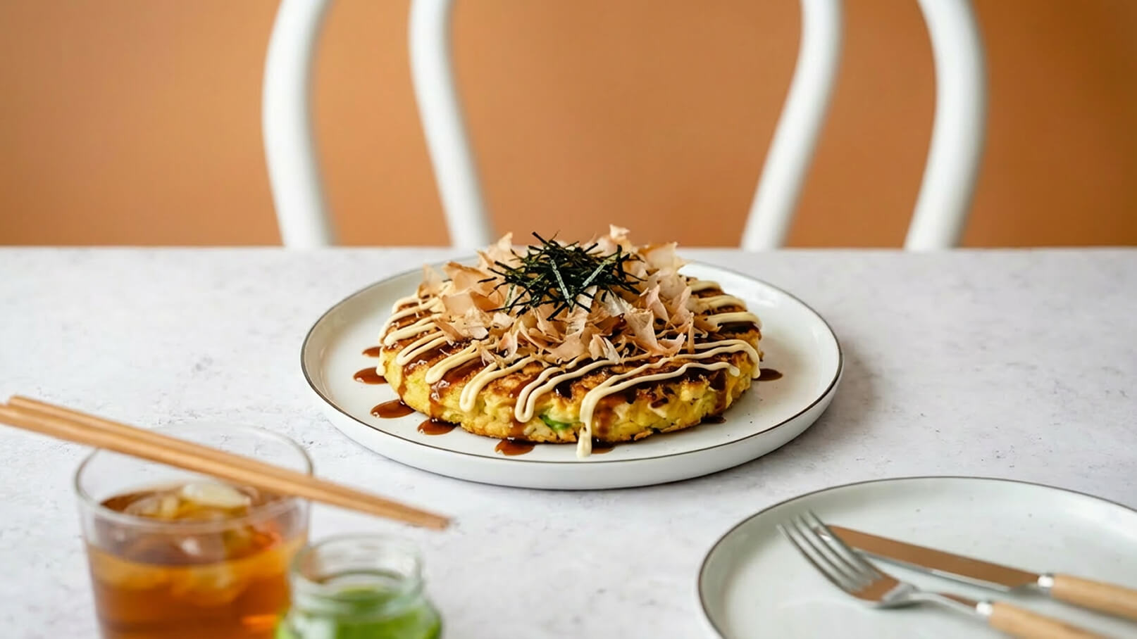 Auf dem Bild ist ein Stück Okonomiyaki, ein japanischer Pfannkuchen, auf einem Teller zu sehen. Er ist mit einem bunten Belag aus Mayonnaise, Soße, getrocknetem Bonito und Nori dekoriert. Im Hintergrund stehen ein Glas Getränk und Essstäbchen bereit.
