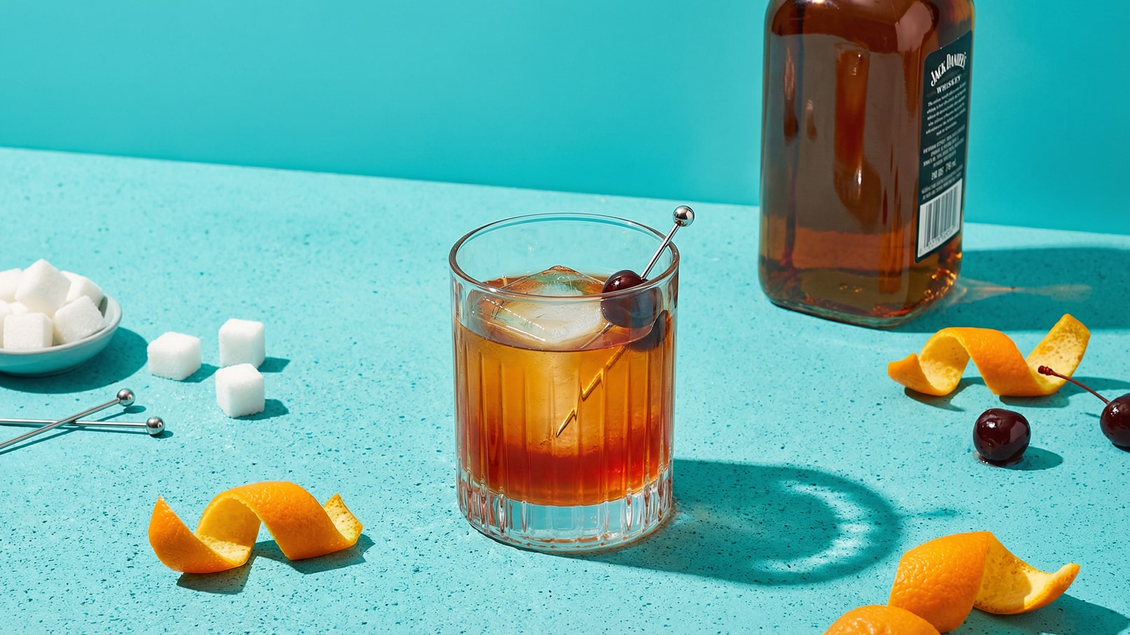 Ein Glas mit einem Drink, gefüllt mit Eiswürfeln und einer Kirsche. Daneben liegen Zuckerwürfel und orangene Schalen. Im Hintergrund steht eine Flasche Whiskey auf einer türkisfarbenen Oberfläche.