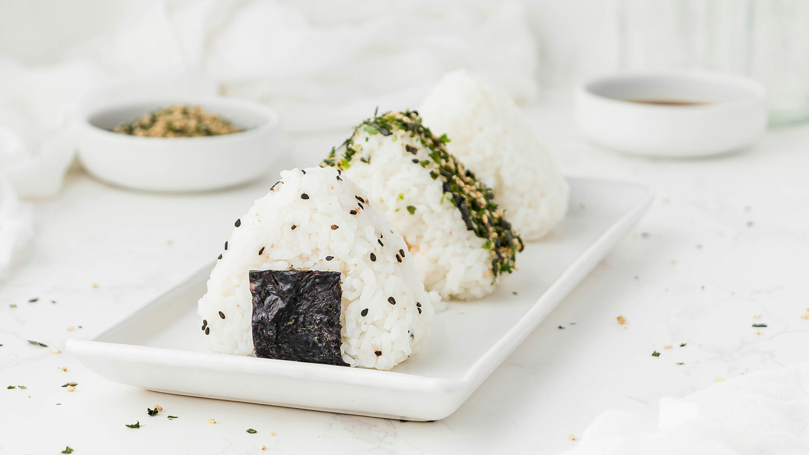 Drei Reisbällchen auf einem weißen Teller, jedes mit unterschiedlicher Garnitur. Eines hat ein schwarzes Nori-Blatt, das andere ist mit grünen Algen und schwarzen Sesamsamen bestreut. Im Hintergrund ist eine kleine Schüssel mit gewürztem Sesam und eine weitere Schüssel mit Sauce sichtbar.