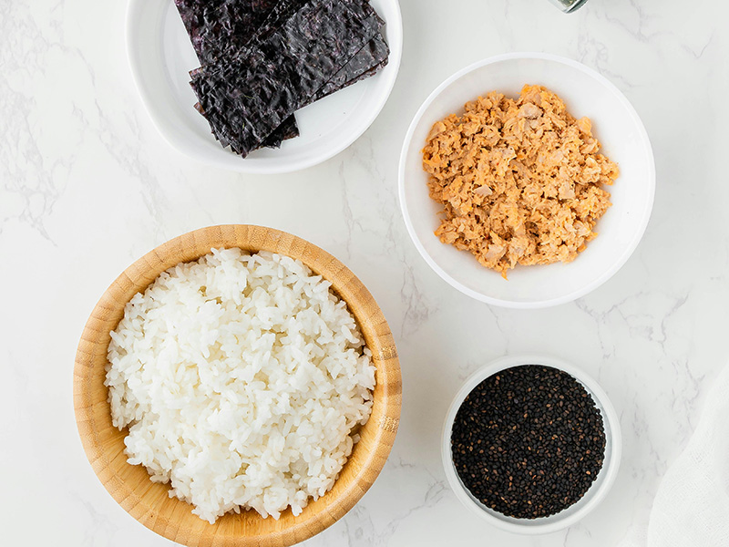Bildbeschreibung: Eine Schüssel mit gekochtem Reis steht im Vordergrund. Neben der Schüssel sind Teller mit zerkleinertem Fisch und geröstetem schwarzen Sesam sowie Blätter aus Nori-Algen angeordnet. Die Zutaten sind auf einer hellen, schlichten Oberfläche platziert.