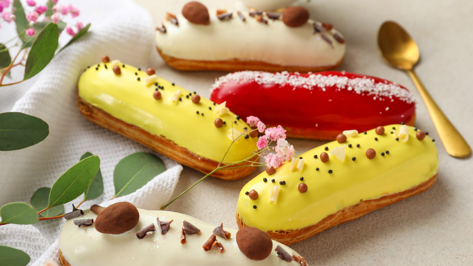 Vier verschiedene Eclairs auf einem hellen Tisch. Die Eclairs sind mit buntem Zuckerguss in Gelb, Rot und Weiß überzogen und mit verschiedenen Dekoren wie Schokoladensplittern und kleinen Pralinen verziert. Einige essbare Blumen und Blätter sind um die Gebäckstücke herum platziert.
