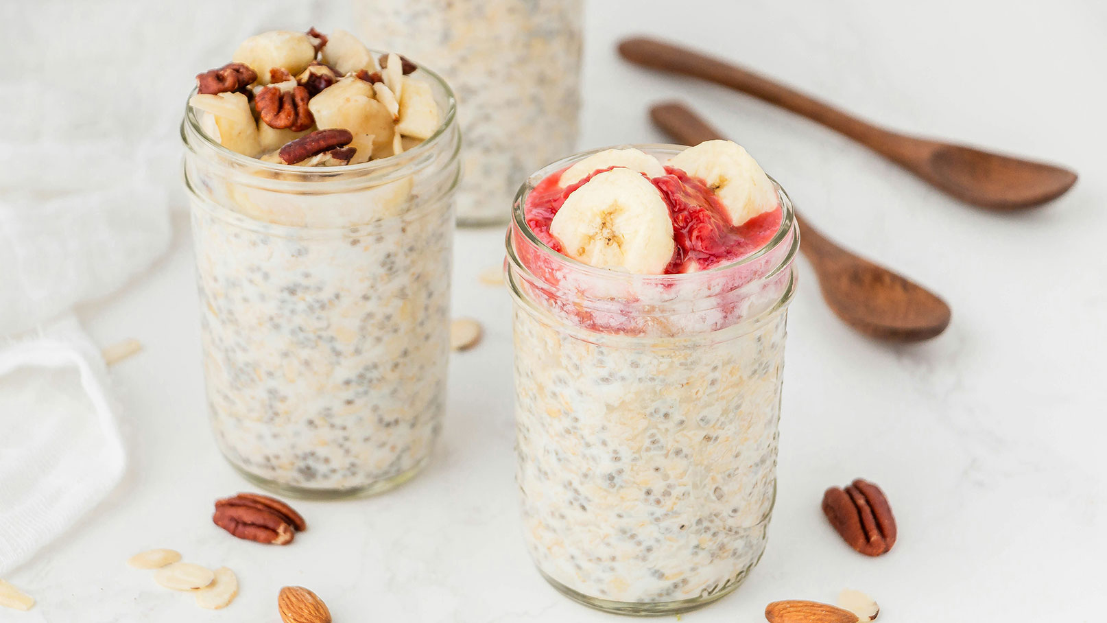 Zwei Gläser mit Overnight Oats stehen auf einer weißen Oberfläche. Das linke Glas ist mit Bananenscheiben und Nüssen garniert, das rechte Glas zeigt glänzende Haferflocken mit einer Schicht aus roter Fruchtsoße und Bananenscheiben.