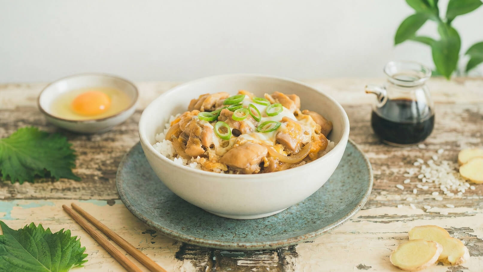 Eine Schüssel mit Reis, belegt mit Hühnchen, Eiern und Frühlingszwiebeln. Daneben ein kleiner Teller mit einem rohen Ei und ein Glas mit einer dunklen Flüssigkeit. Die Szenerie ist auf einem rustikalen Tisch angeordnet.