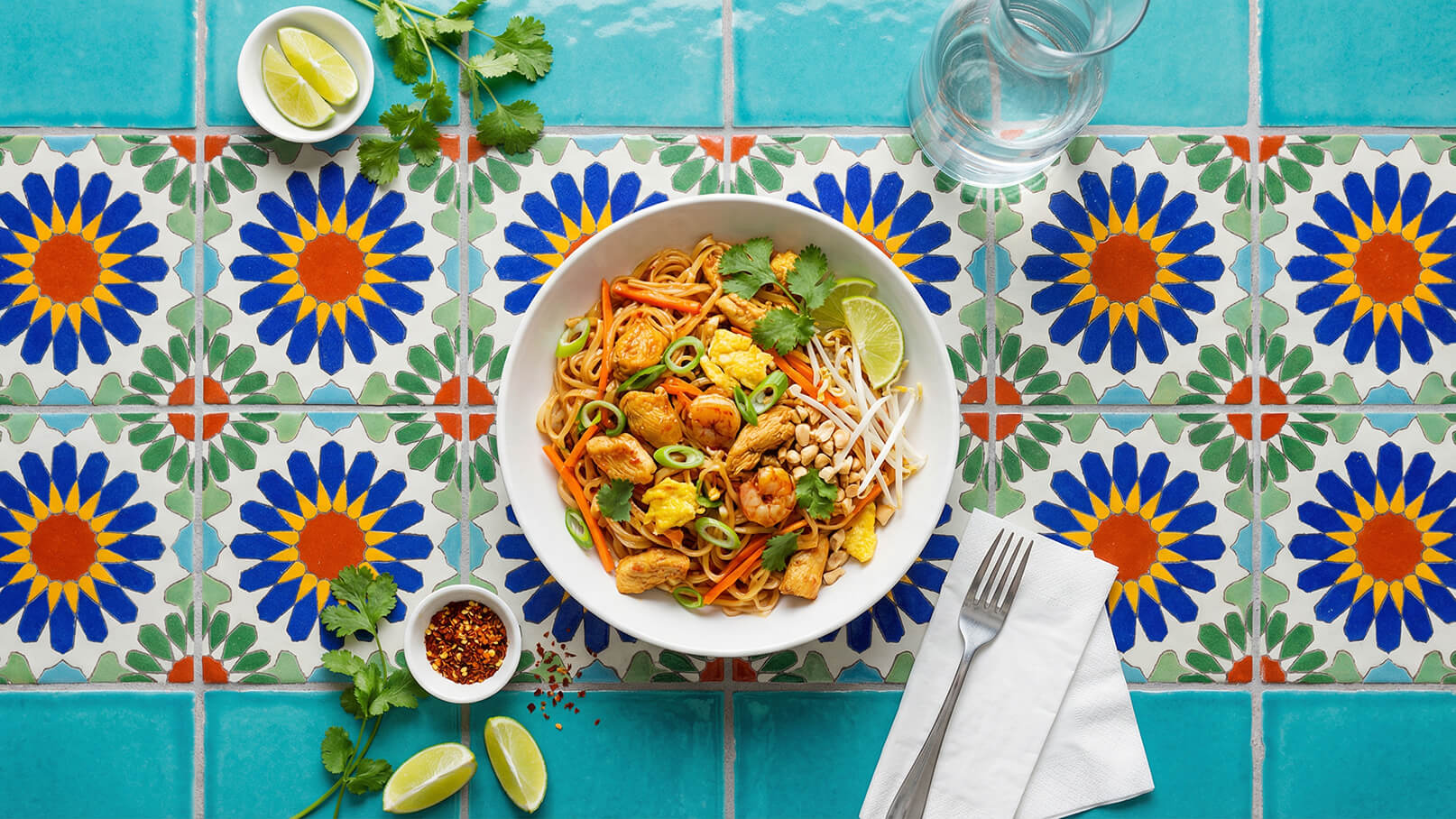 Eine Schüssel mit gebratenen Nudeln, Garnelen, Hühnchen, Gemüse und frischen Kräutern steht auf einem bunten, blumenmusternden Tisch. Neben der Schüssel liegen eine Gabel und Servietten. Im Hintergrund befinden sich Limetten und ein Glas Wasser.