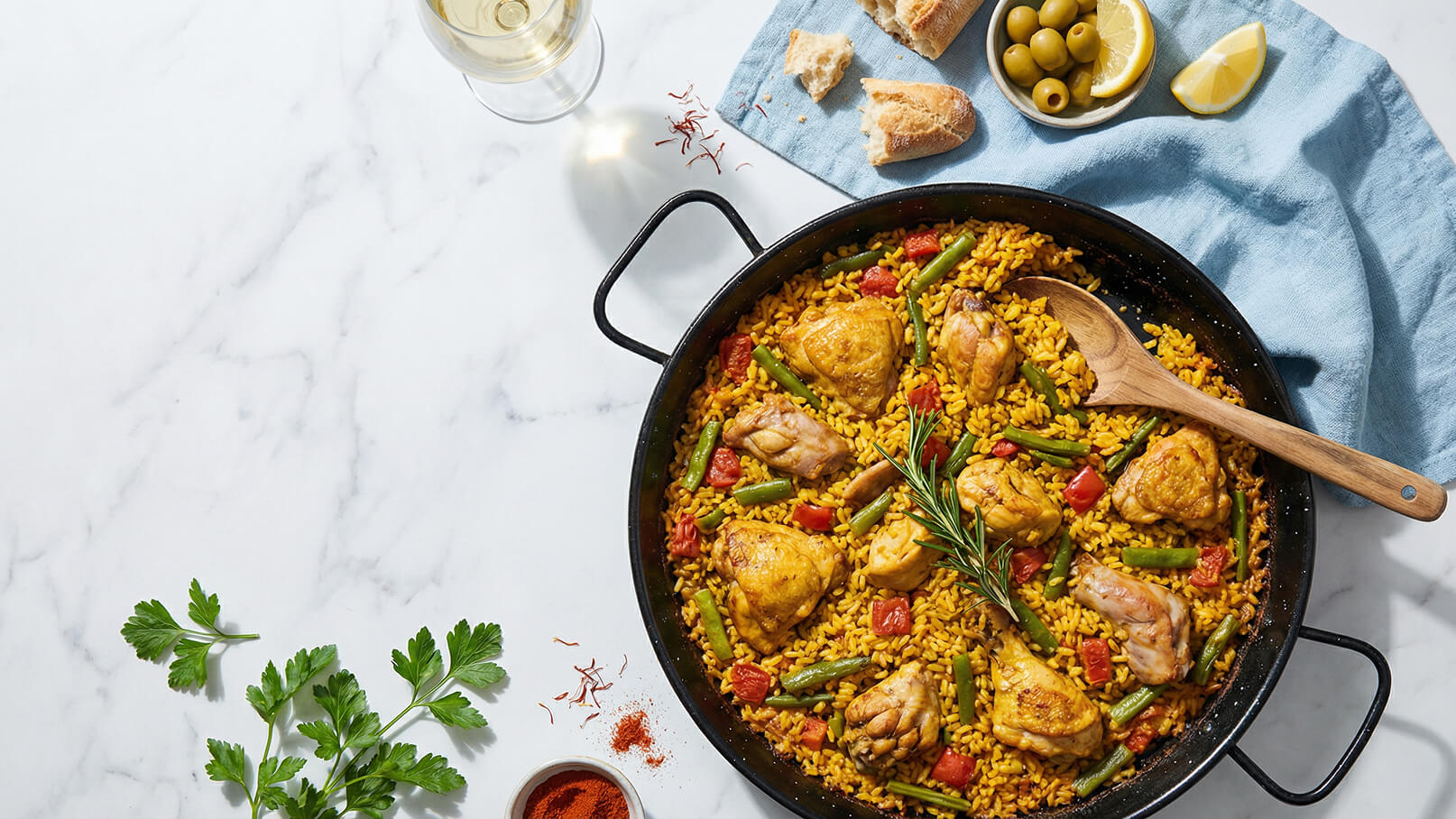 Eine große Pfanne mit gelbem Reis, Hähnchenteilen, grünen Bohnen und Tomaten. Ein Holzlöffel liegt neben dem Gericht. Daneben liegen frisches Brot, grüne Oliven und Zitronenscheiben auf einem blauen Tuch.