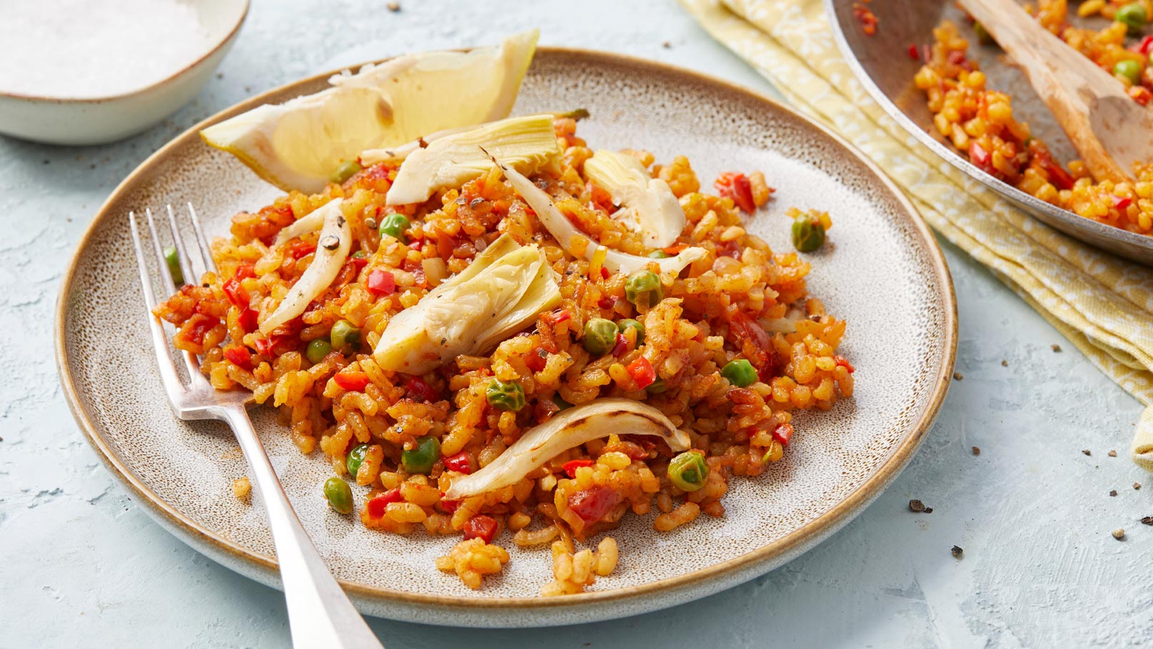 Ein Teller mit buntem Reisgericht, das mit Paprika, Erbsen und Artischockenstücken garniert ist. Eine Gabel liegt neben dem Gericht. Im Hintergrund ist eine Schüssel mit Salz und eine Serviette sichtbar.