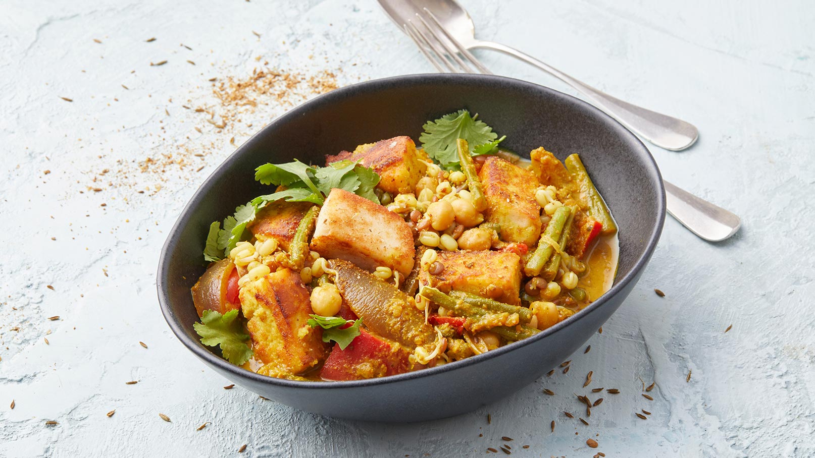 Schüssel mit buntem Gemüse, Kichererbsen, und gewürztem Tofu. Das Gericht ist mit frischem Koriander garniert und auf einem hellen Untergrund angerichtet. Es vermittelt einen Eindruck von herzhaftem, vegetarischem Essen.