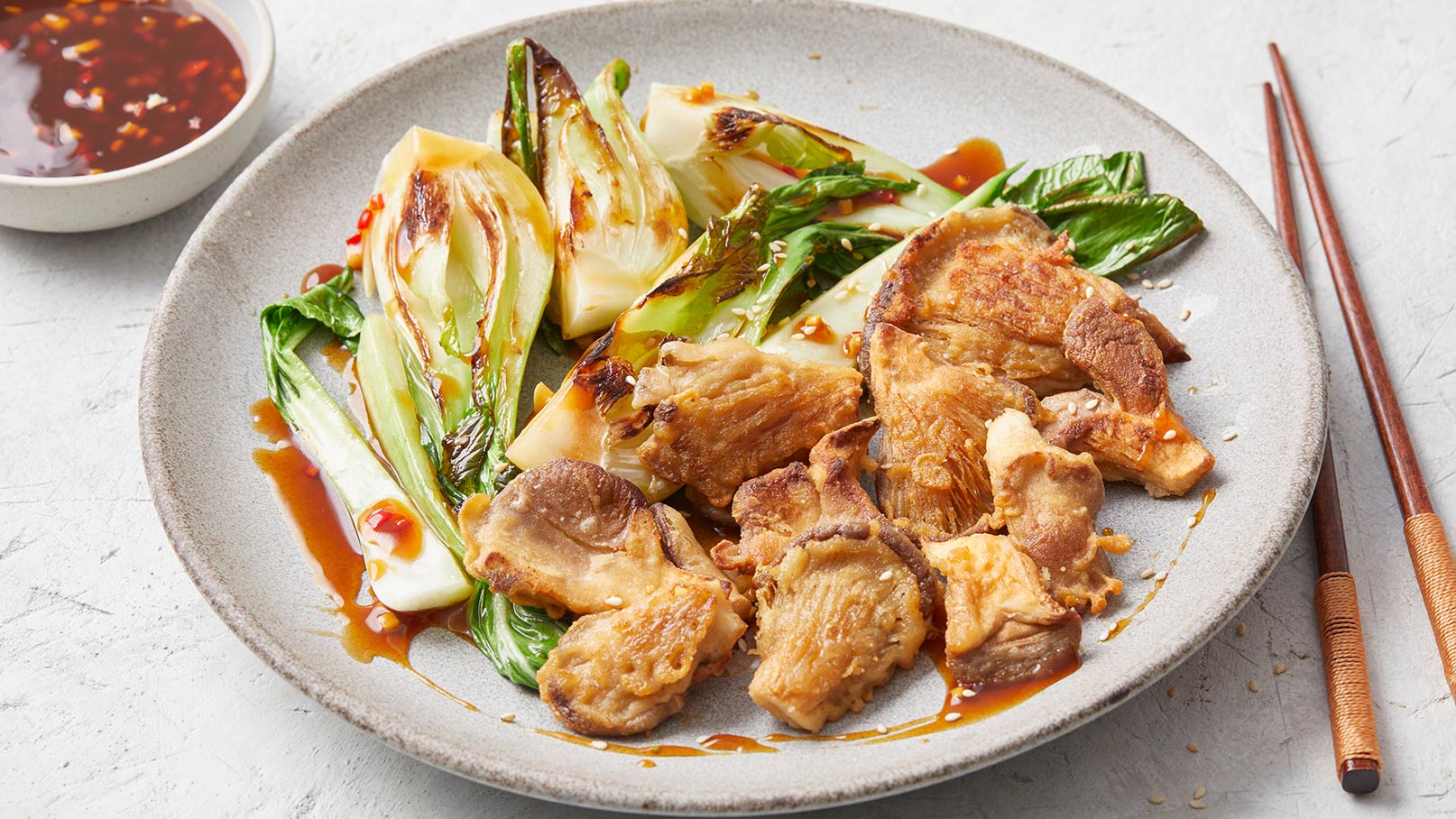 Ein Teller mit gebratenem Fleischstücken und sautiertem Pak Choi, serviert mit einer dunklen Soße. Neben dem Teller befinden sich Essstäbchen.