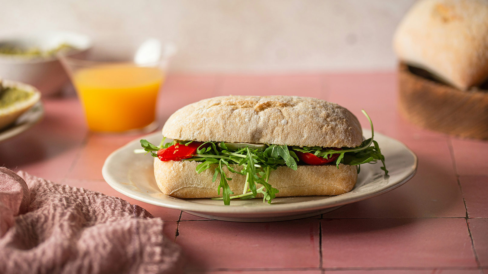 Ein belegtes Brötchen liegt auf einem Teller. Es ist mit frischem Rucola und roten Tomatenscheiben gefüllt. Im Hintergrund ist ein Glas mit orangem Getränk sichtbar. Der Tisch hat eine rosa Oberfläche.