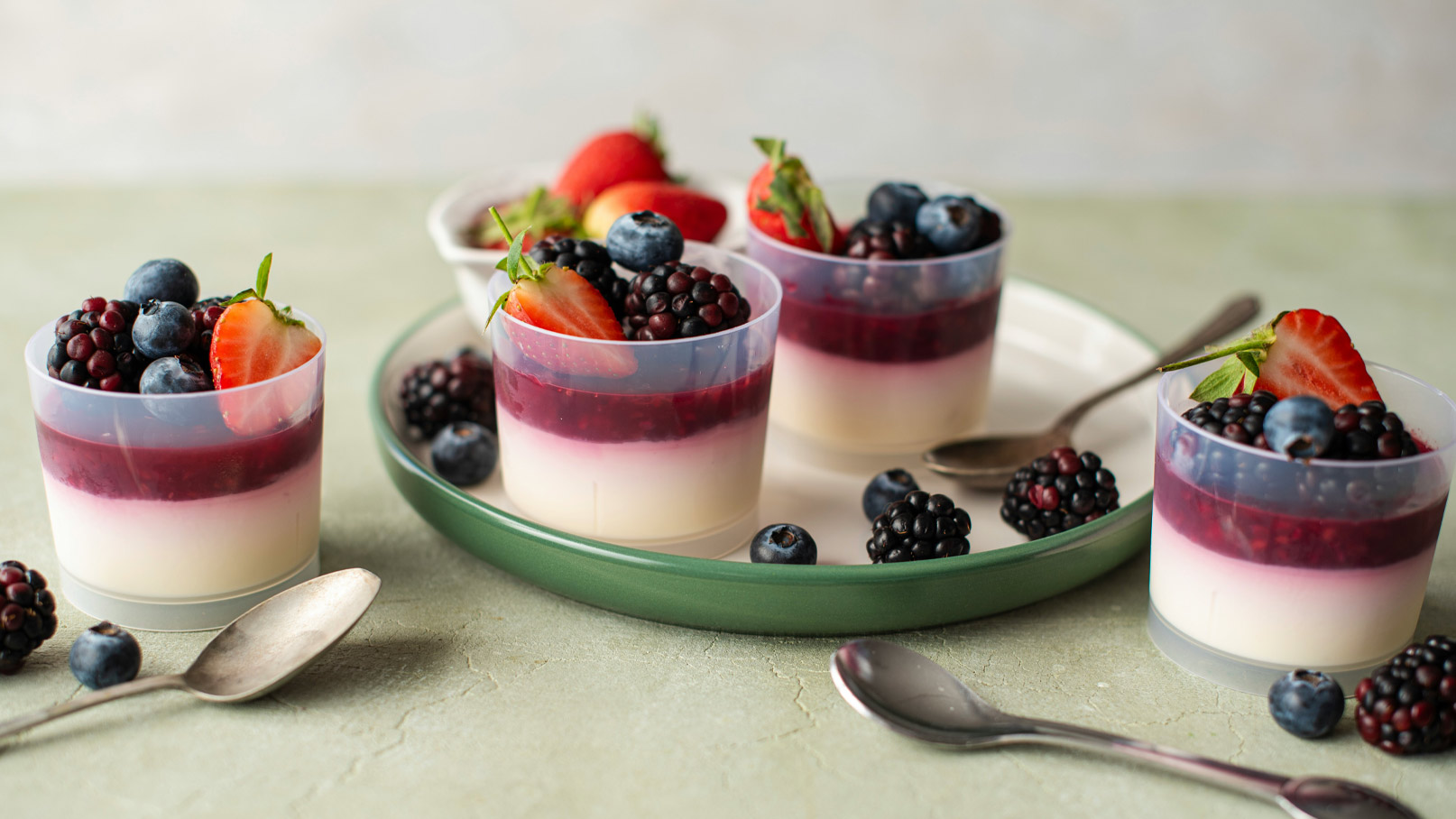 Vier transparente Becher mit einem Dessert, bestehend aus einer Schicht vanilliger Creme und einer Beerensoße obendrauf, garniert mit frischen Erdbeeren, Heidelbeeren und Brombeeren. Im Hintergrund steht ein grüner Teller mit weiteren Beeren und einem Löffel.