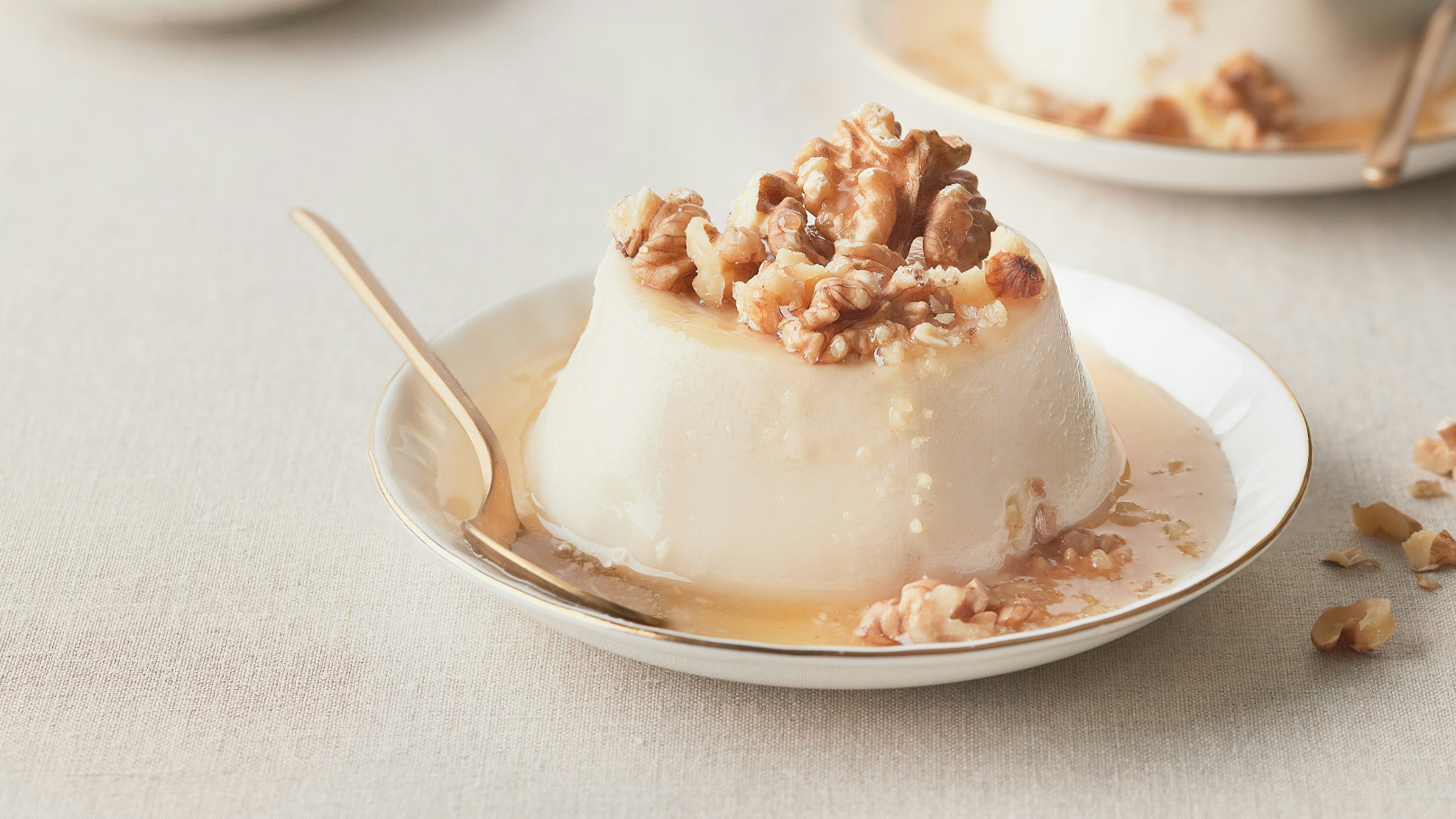 Ein Dessert auf einem weißen Teller: eine samtige Panna Cotta, garniert mit gehackten Walnüssen und einem süßen Sirup. Eine Gabel liegt neben dem Teller. Der Hintergrund ist hell und neutral gehalten.
