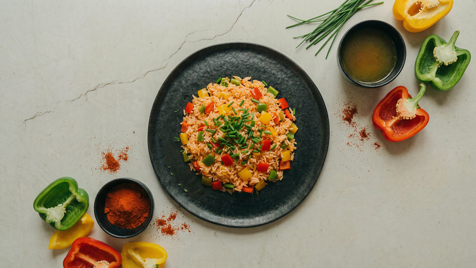 Ein schwarzer Teller mit buntem Reis, garniert mit frischem Schnittlauch und verschiedenen Paprikawürfeln in Rot, Grün und Gelb. Daneben befinden sich geschnittene Paprika und eine kleine Schale mit Gewürzpulver.