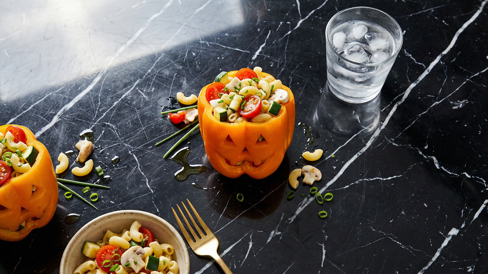 Ein orangefarbener Paprika, der wie ein Kürbis geschnitzt ist, ist mit einer Mischung aus Pasta, Kirschtomaten, Zucchini und Frühlingszwiebeln gefüllt. Daneben liegt ein kleiner Teller mit ähnlicher Pasta und ein Glas Wasser. Der Hintergrund ist schwarz marmoriert.