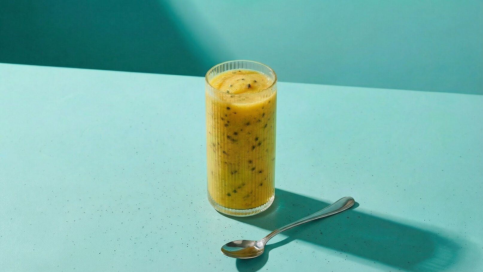 Ein hohes Glas mit einem gelben, fruchtigen Smoothie steht auf einer blauen Oberfläche. Der Smoothie hat eine cremige Textur und kleine Fruchtstücke, während ein Löffel daneben liegt. Das Licht erzeugt sanfte Schatten und betont die Farben.