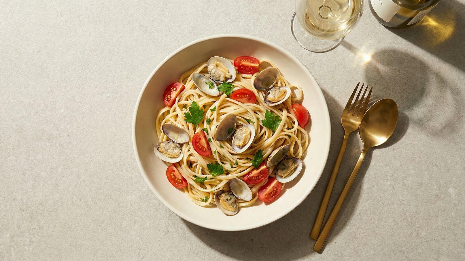 Ein tiefer Teller mit Spaghetti, garniert mit Muscheln, Kirschtomaten und frischer Petersilie. Daneben liegen goldenes Besteck und ein Glas Wein. Der Teller ist auf eine helle Oberfläche gesetzt.