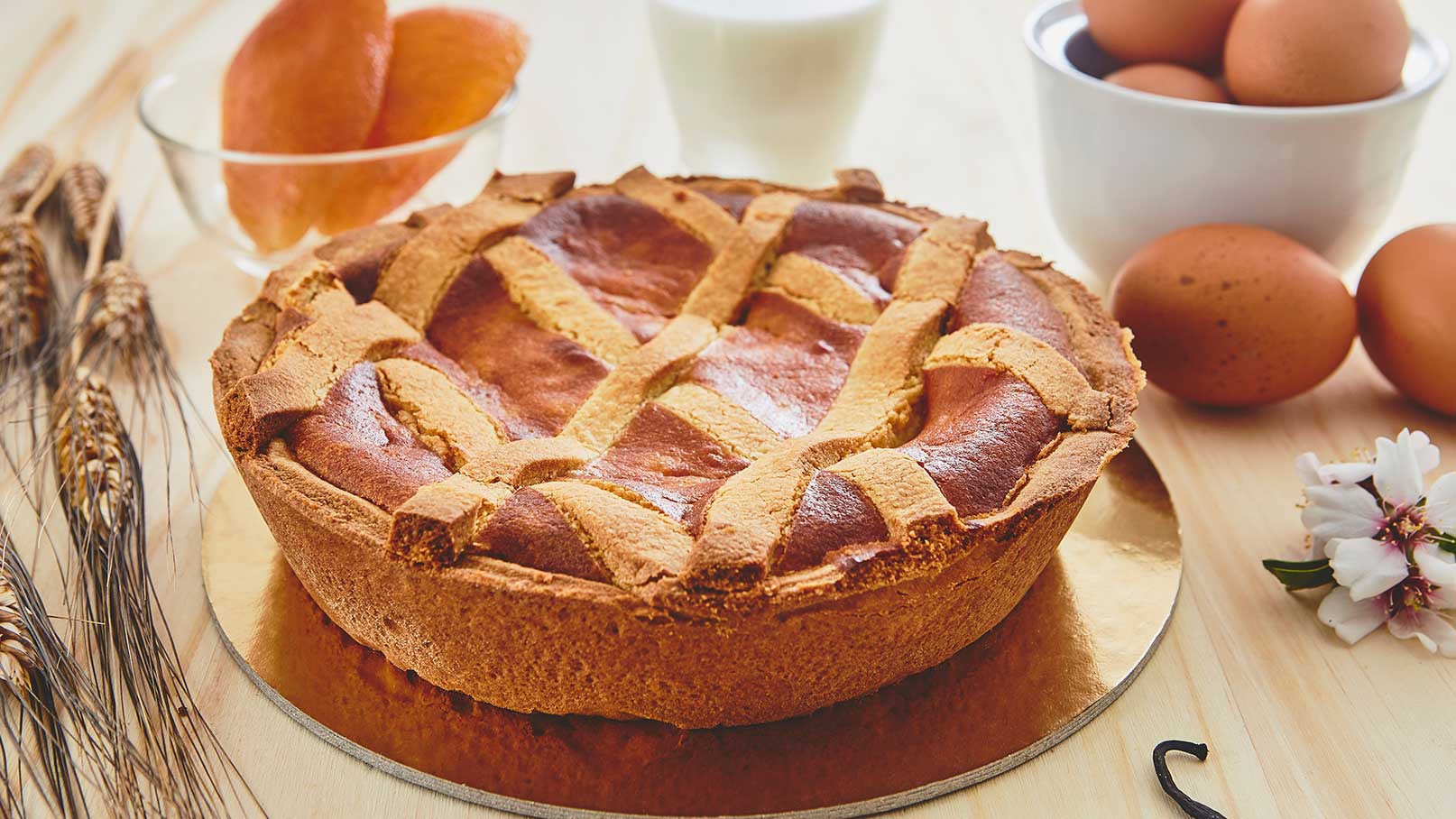 Ein frisch gebackener Kuchen mit einem geflochtenen Teiggitter ist auf einer runden Unterlage platziert. Im Hintergrund sind eine Schüssel mit Eiern, ein Glas Milch und eine Schüssel mit Früchten zu sehen. Die Holzoberfläche vermittelt eine gemütliche Küchenszenen.