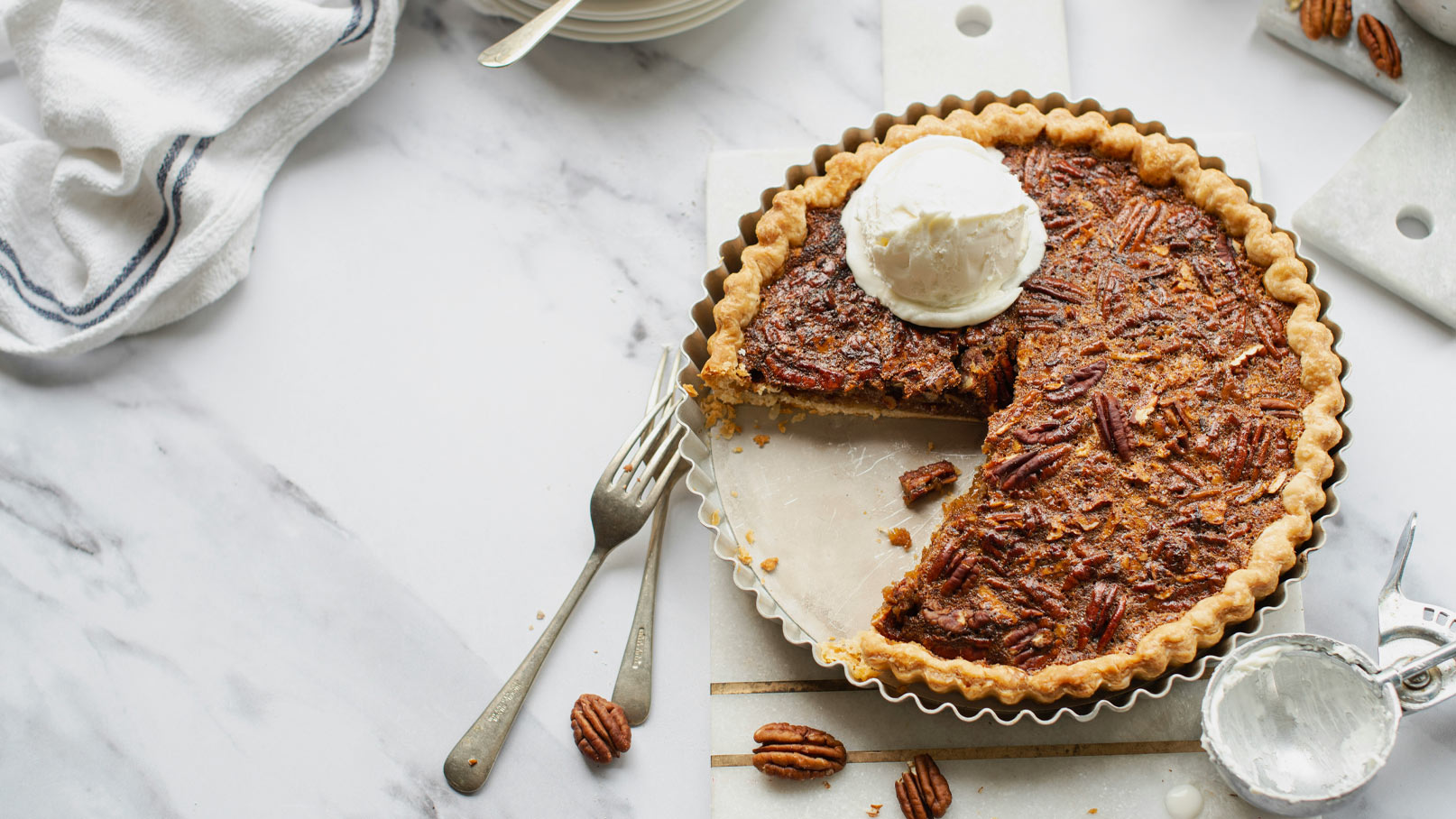 Ein köstlicher Pecan-Pie mit einer Portion Vanilleeis auf der Oberseite. Ein Stück ist bereits entnommen, und die Torte steht auf einer hellen Marmortheke. Einige Pekannüsse sind um die Torte verstreut.