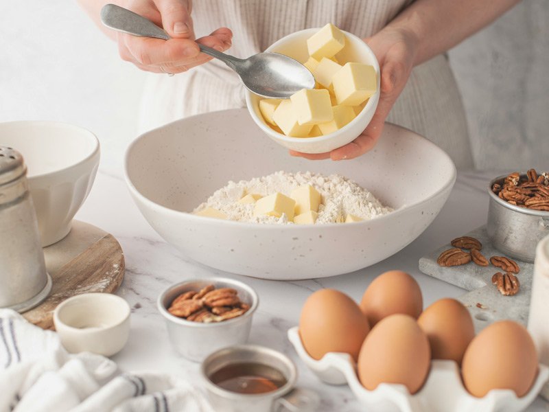 Eine Hand hält eine Schüssel mit Butterstücken und einen Löffel, um die Butter in eine Schüssel mit Mehl zu geben. Im Vordergrund sind Eier und eine kleine Schüssel mit Nüssen sichtbar. Die Szene zeigt Vorbereitungen für das Backen.