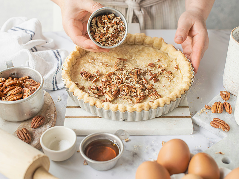 Person gibt Nüsse zu einem Kuchen auf einer Küchenarbeitsplatte, umgeben von Backzutaten, darunter Eier und Gewürzschalen.