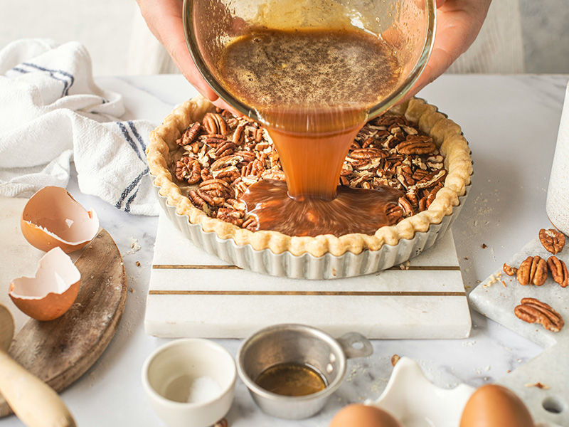 Eine Person gießt eine Karamellmischung in den Boden einer Pekannuss-Torte. Um die Torte herum liegen Eierschalen, Mehl, Pekannüsse.