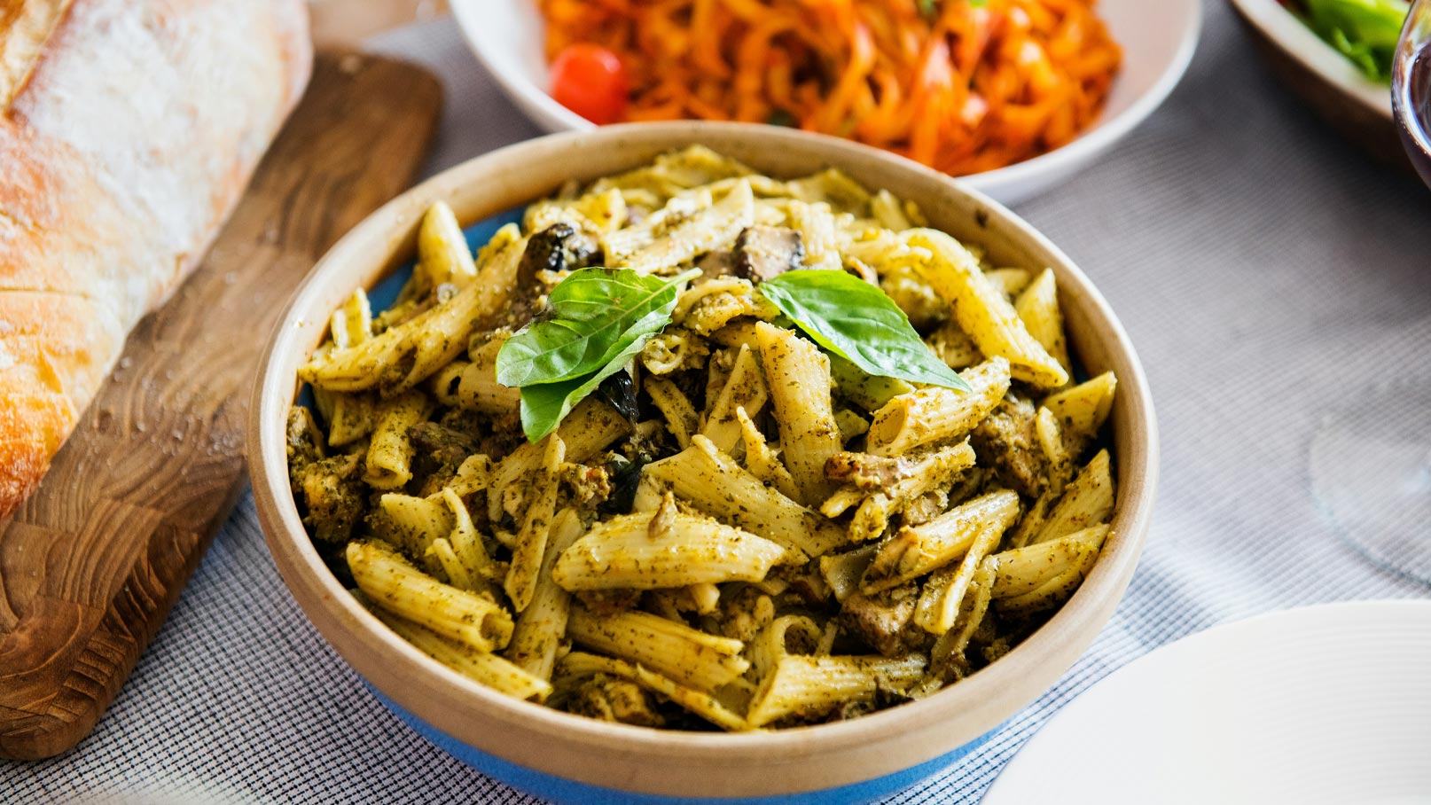 Ein brauner Schüssel mit Penne-Pasta ist mit grüner Basilikum- und Pesto-Sauce garniert. Im Hintergrund sind bunt angerichtete Beilagen zu sehen. Der Tisch ist mit einem grauen Tuch gedeckt, während ein Laib Brot auf einem Holzbrett liegt.