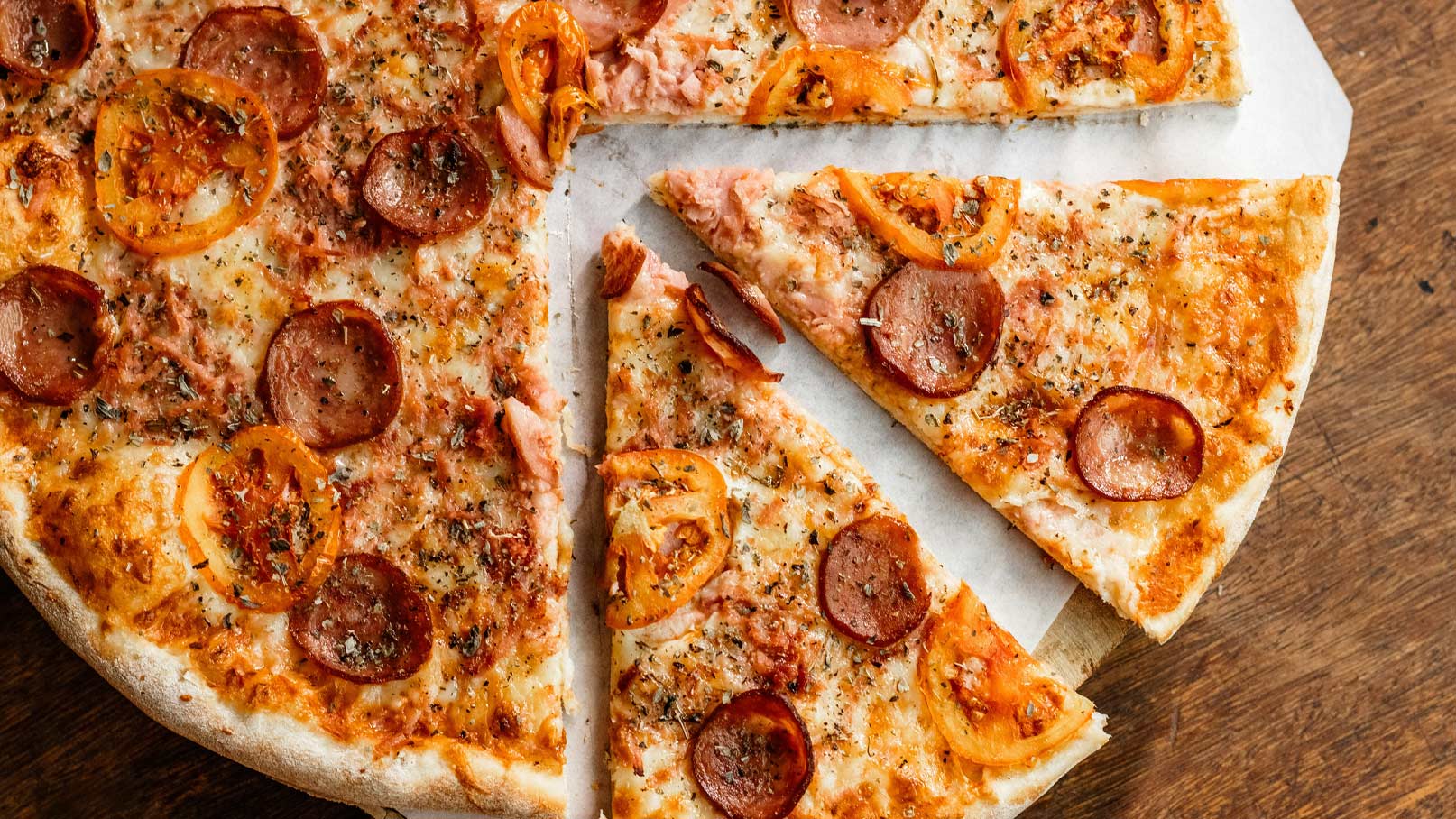 Eine Pizza mit goldbraunem Käse, belegt mit Salami und roten Paprikastücken. Einige Stücke sind bereits abgeschnitten und platziert, während andere auf der runden Pizza bleiben. Der Boden ist knusprig und die Zutaten sind gleichmäßig darauf verteilt.