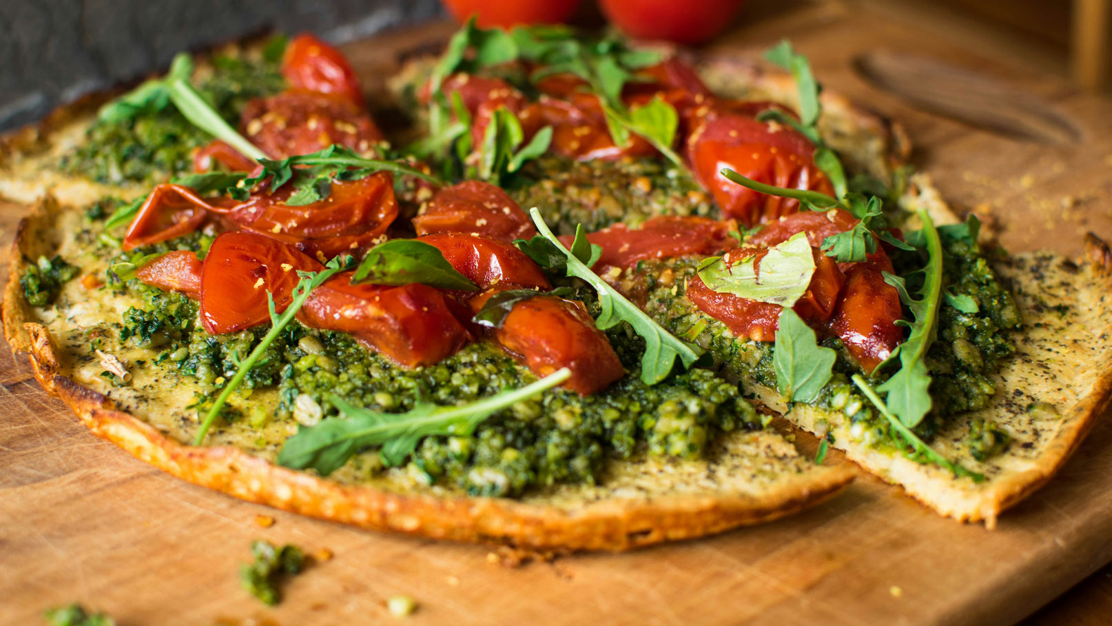 Ein frisch zubereiteter Flammkuchen, belegt mit grüner Pesto-Sauce, saftigen Tomatenstücken und frischem Rucola. Der Teig ist goldbraun und knusprig. Der Flammkuchen liegt auf einem Holzbrett, umgeben von Tomaten und Kräutern.