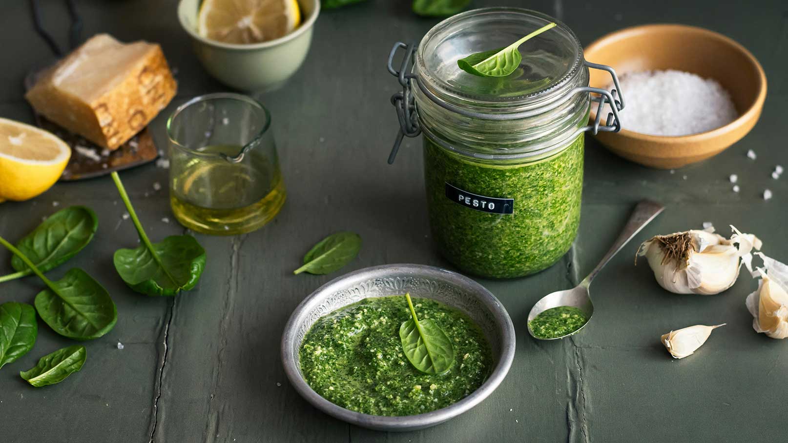 Ein Glas mit grünem Pesto steht auf einem dunklen Tisch. Daneben befindet sich eine kleine Schüssel mit Pesto und frischen Spinatblättern. Weitere Zutaten sind eine Zitrone, ein Glas Olivenöl und Knoblauchzehen. Das Bild vermittelt eine frische, geschmackvolle Kochatmosphäre.
